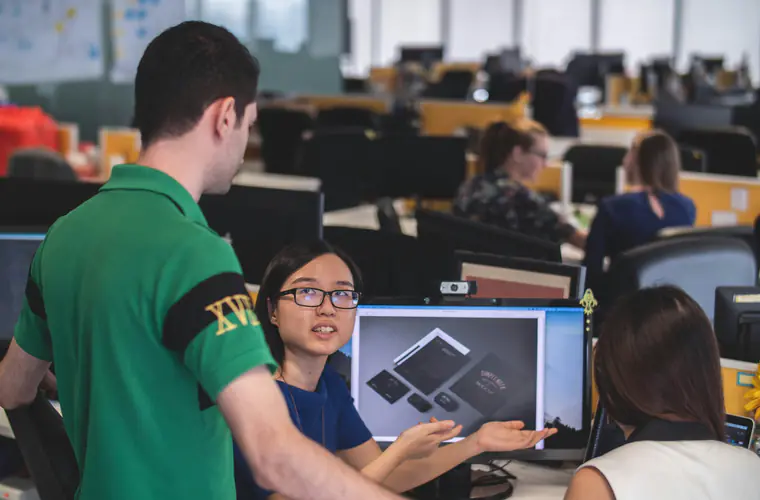 Colleagues gathered around a monitor in an open-plan office, working through something on screen together.