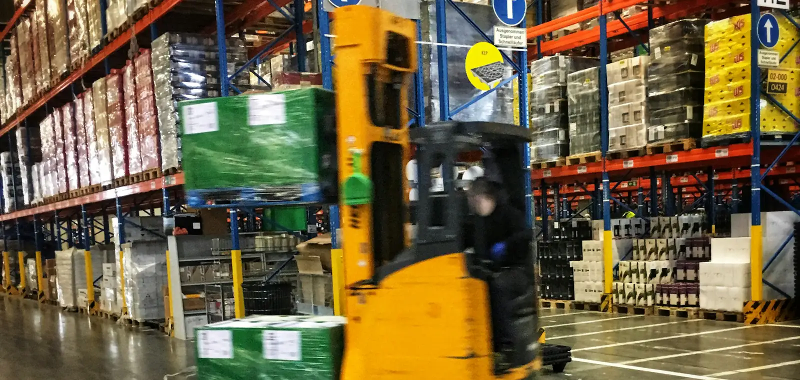 Reach truck moving a pallet down a lit warehouse aisle, racking stacked several levels high on both sides.