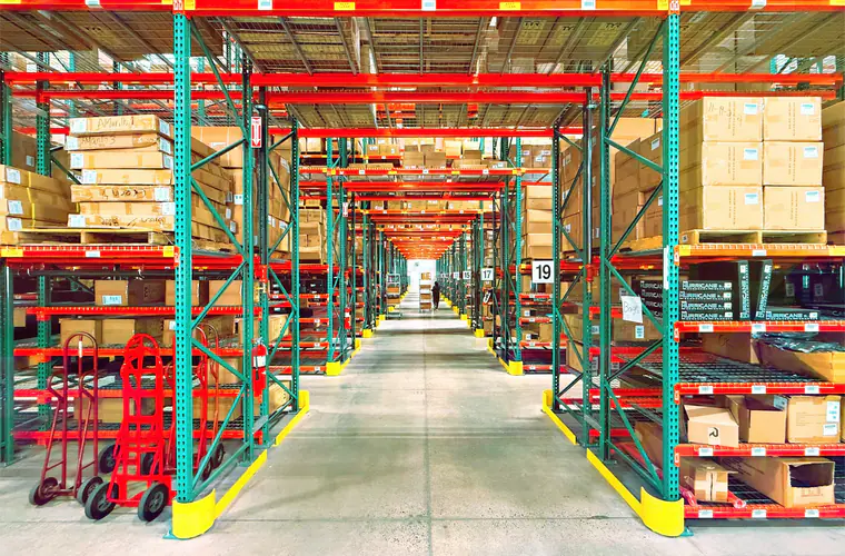 Racking aisle in a distribution warehouse stacked with cartons, hand trucks parked at the end of the row.